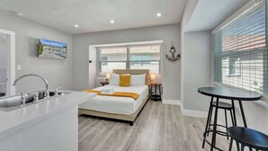 A modern bedroom with a bed, bedside tables, a window with blinds, and a small kitchen area with a sink and countertop. - Walk to Beach Studio | Pompano Beach (Pompano Beach)