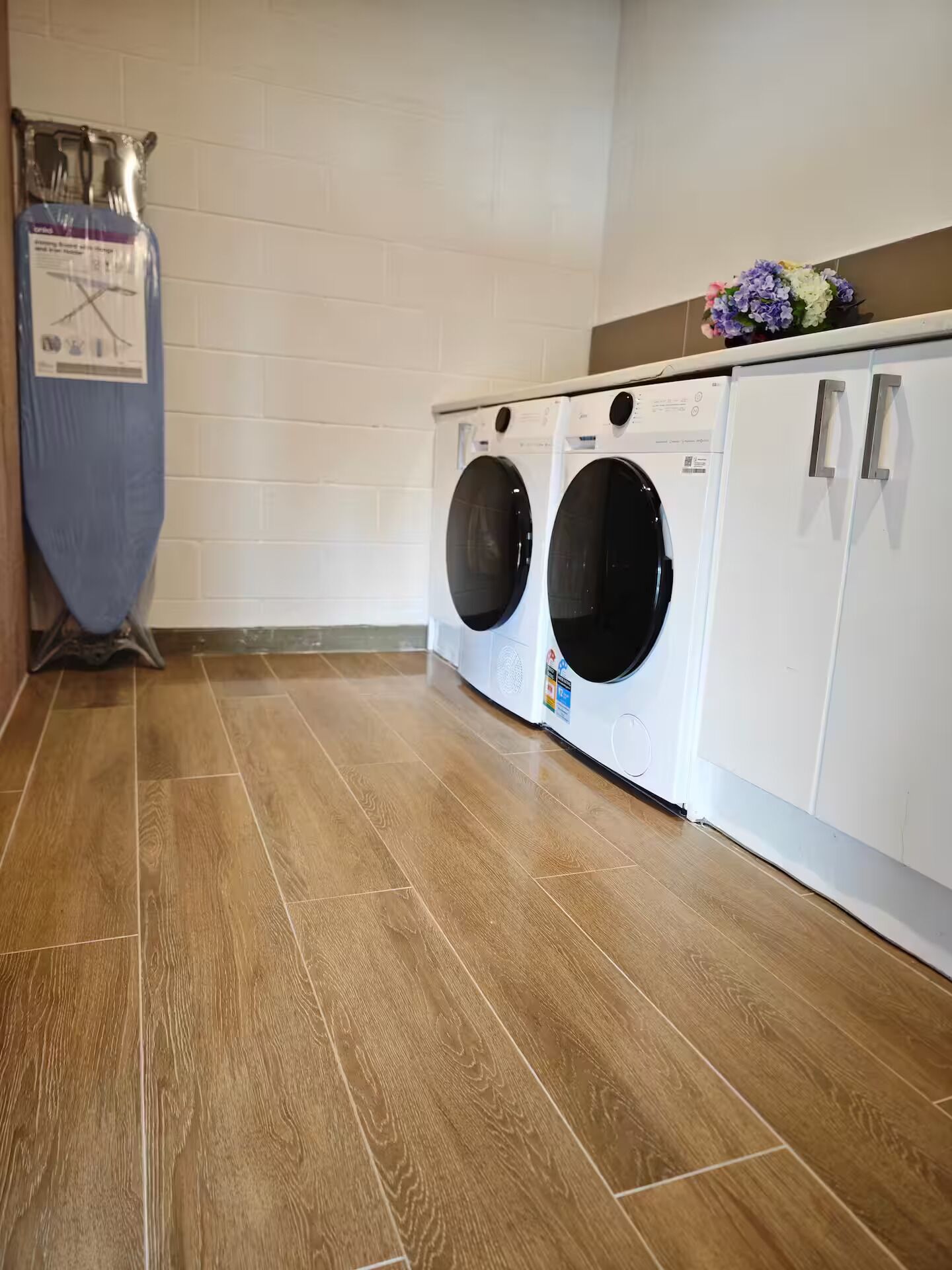 A laundry room with two front-loading washing machines, a drying rack, and a cabinet with a floral arrangement.