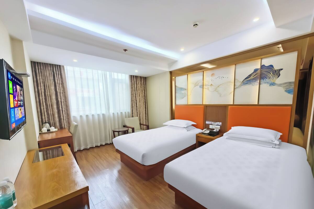 A hotel room with two beds, a desk, a TV, and a window with curtains.