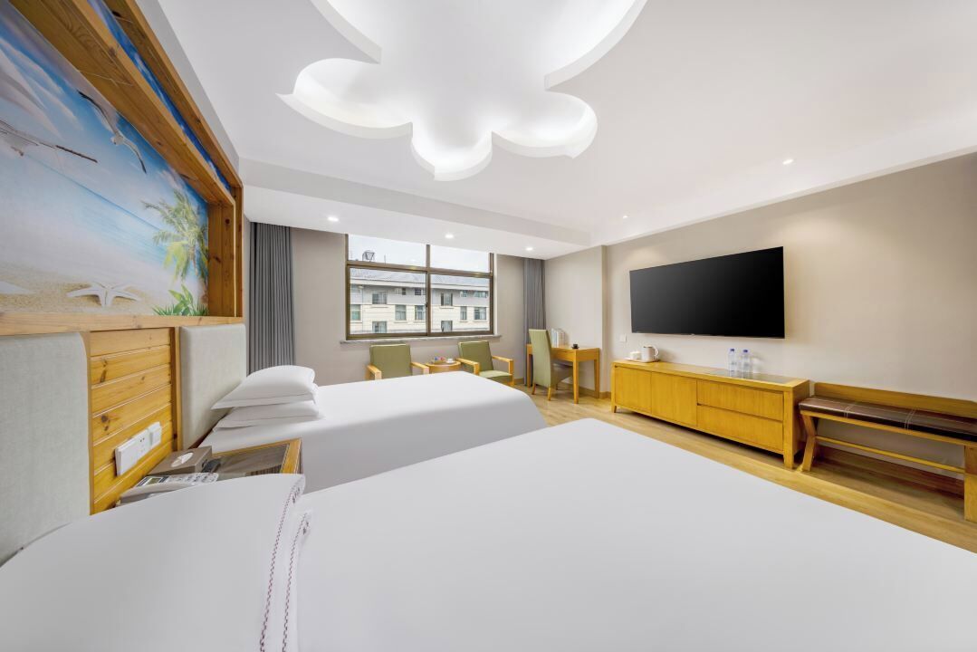 A hotel room with two beds, a TV, a wooden dresser, and a large window with a view of buildings.