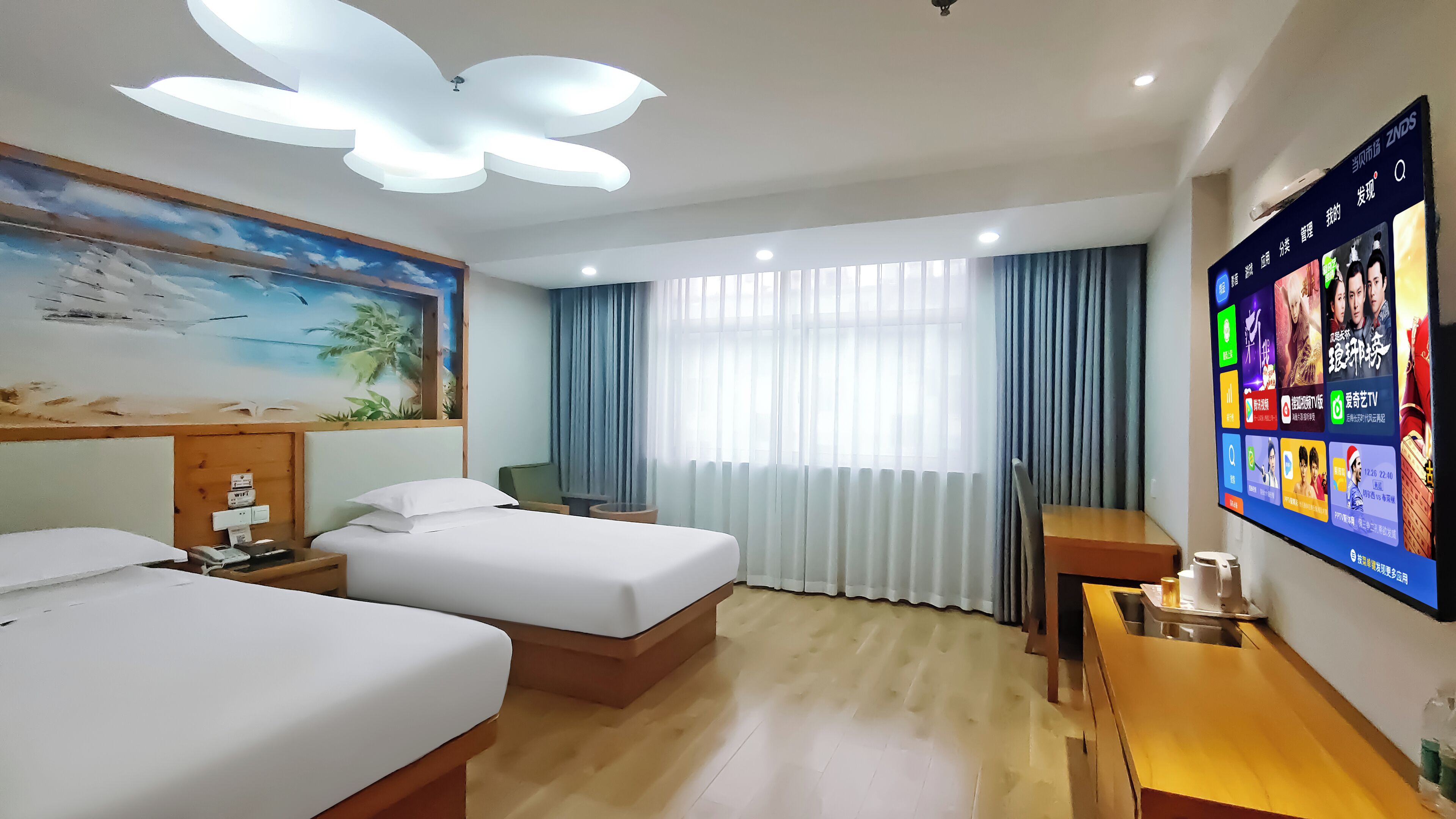 A hotel room with two beds, a desk, a chair, a TV displaying various channels, and a painting of a beach scene.
