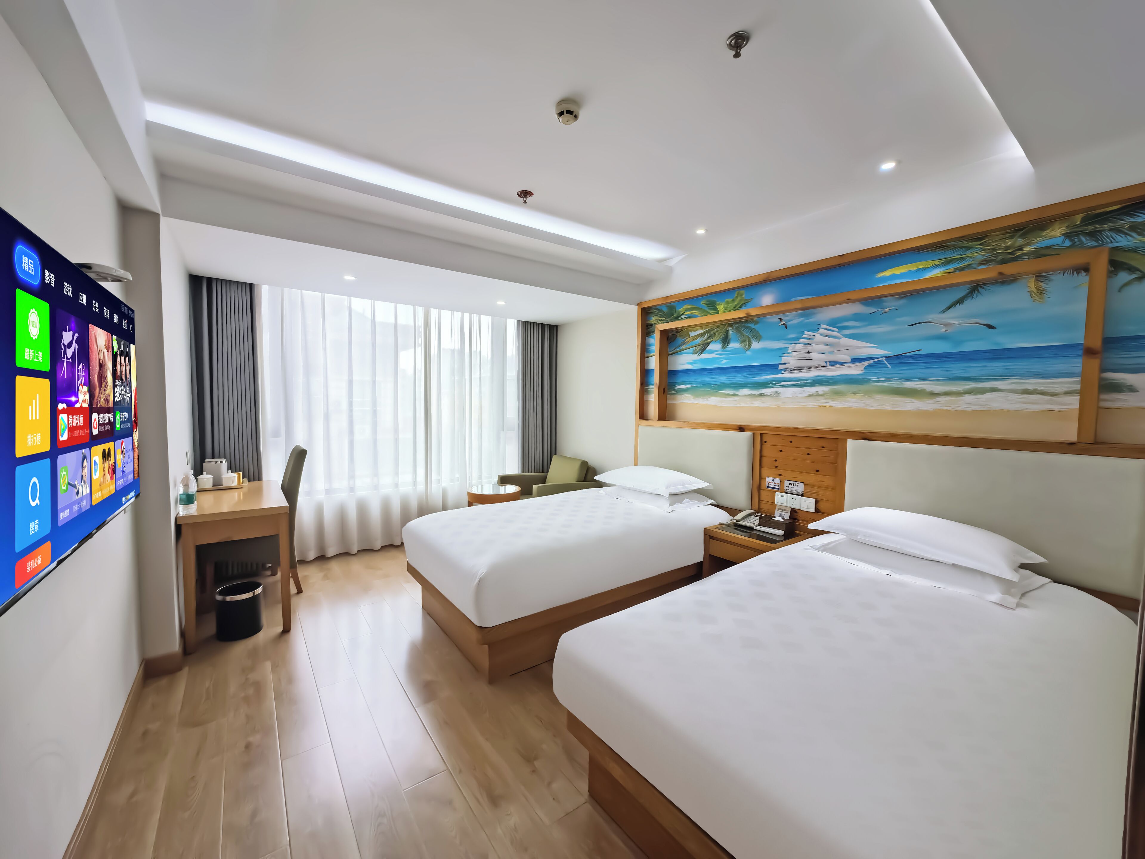 A hotel room with two beds, a desk, a chair, a TV screen, and a painting of a beach scene.