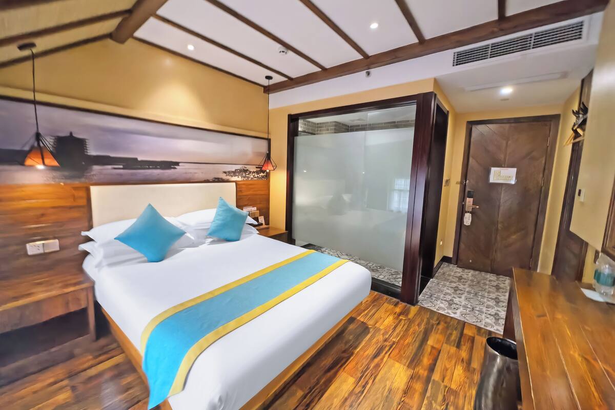 A hotel room with a bed, wooden flooring, a nightstand, a wall-mounted lamp, and a door with a frosted glass panel.