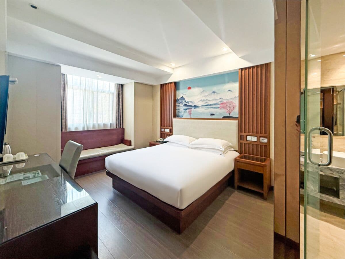 Habitación de hotel con una cama grande, un escritorio, una silla, una mesita, una mesita de noche, un cuadro y un box de ducha de vidrio.