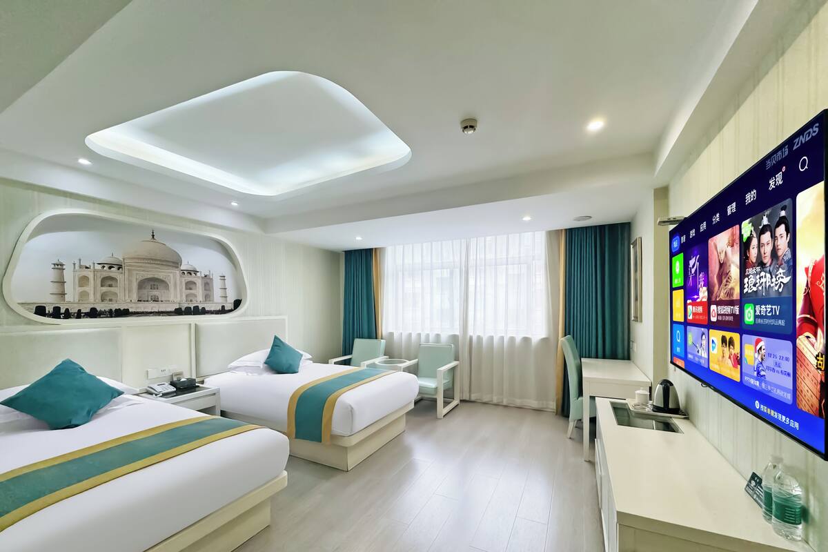 A hotel room with two beds, a TV displaying entertainment options, a desk, and a view of a building through a window.