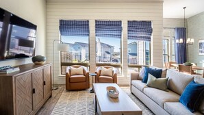 Un salon moderne comprenant un canapé, deux fauteuils, une table basse, un téléviseur et de grandes fenêtres avec des rideaux.