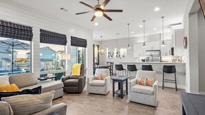 A modern living room with a sofa, armchairs, and a dining table. There is a kitchen area with a refrigerator and a dining table. The room has large windows and a ceiling fan. - The Sandbar Retreat! (Kingsland)