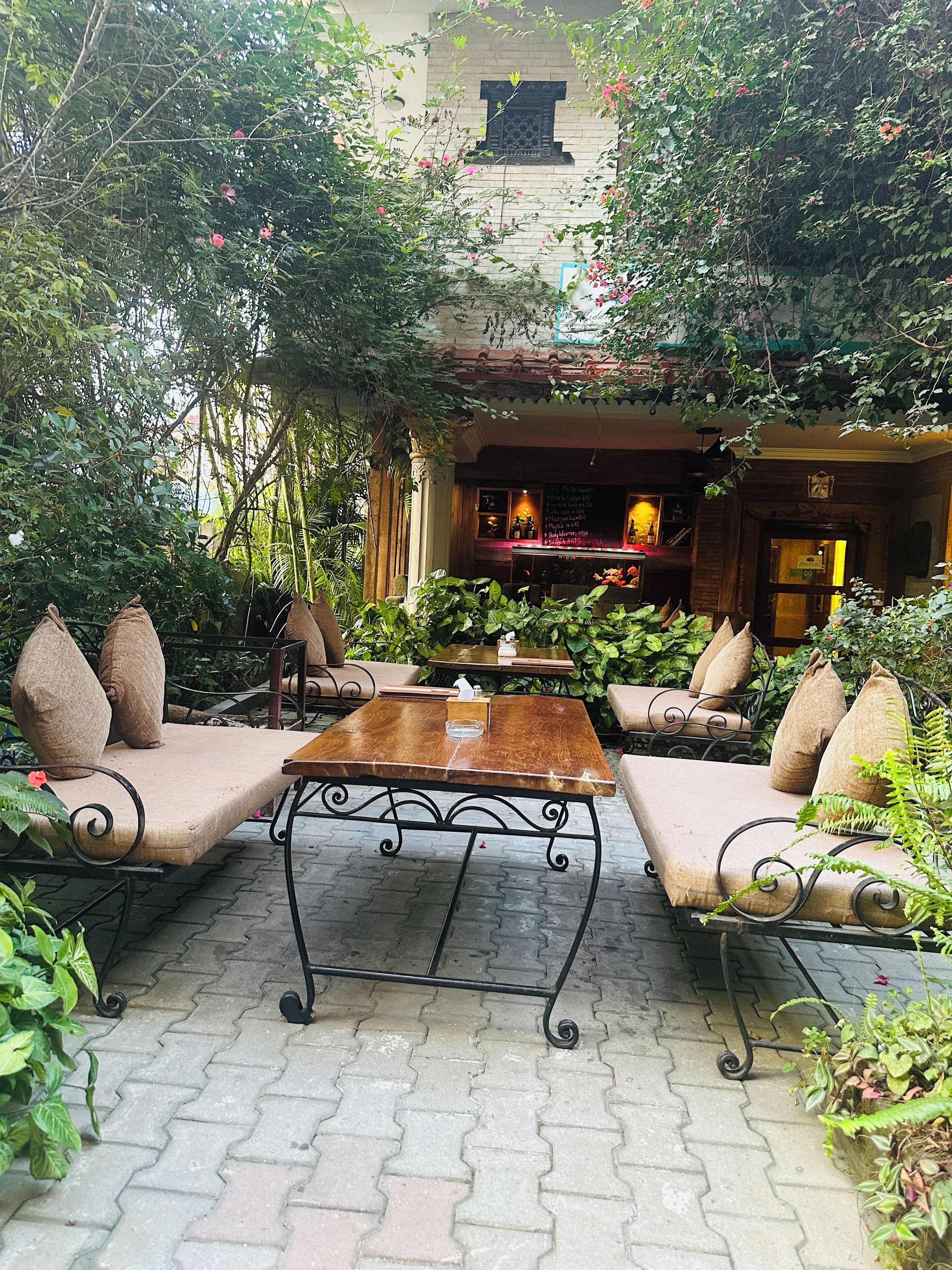 An outdoor seating area with cushioned sofas and a wooden table, surrounded by lush greenery and a brick building in the background.