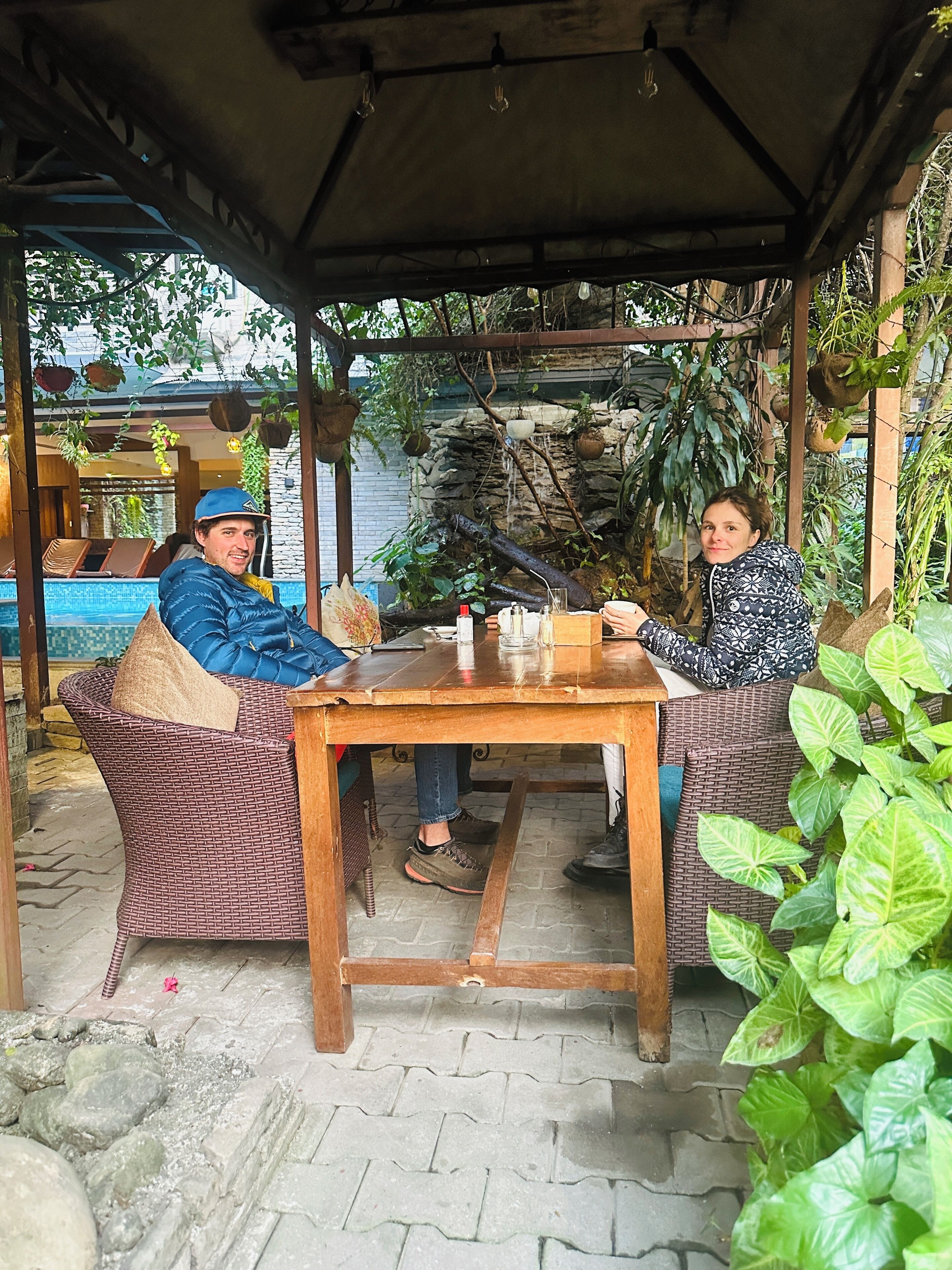 Two people seated at a wooden table under a covered area with hanging plants and a stone wall in the background.