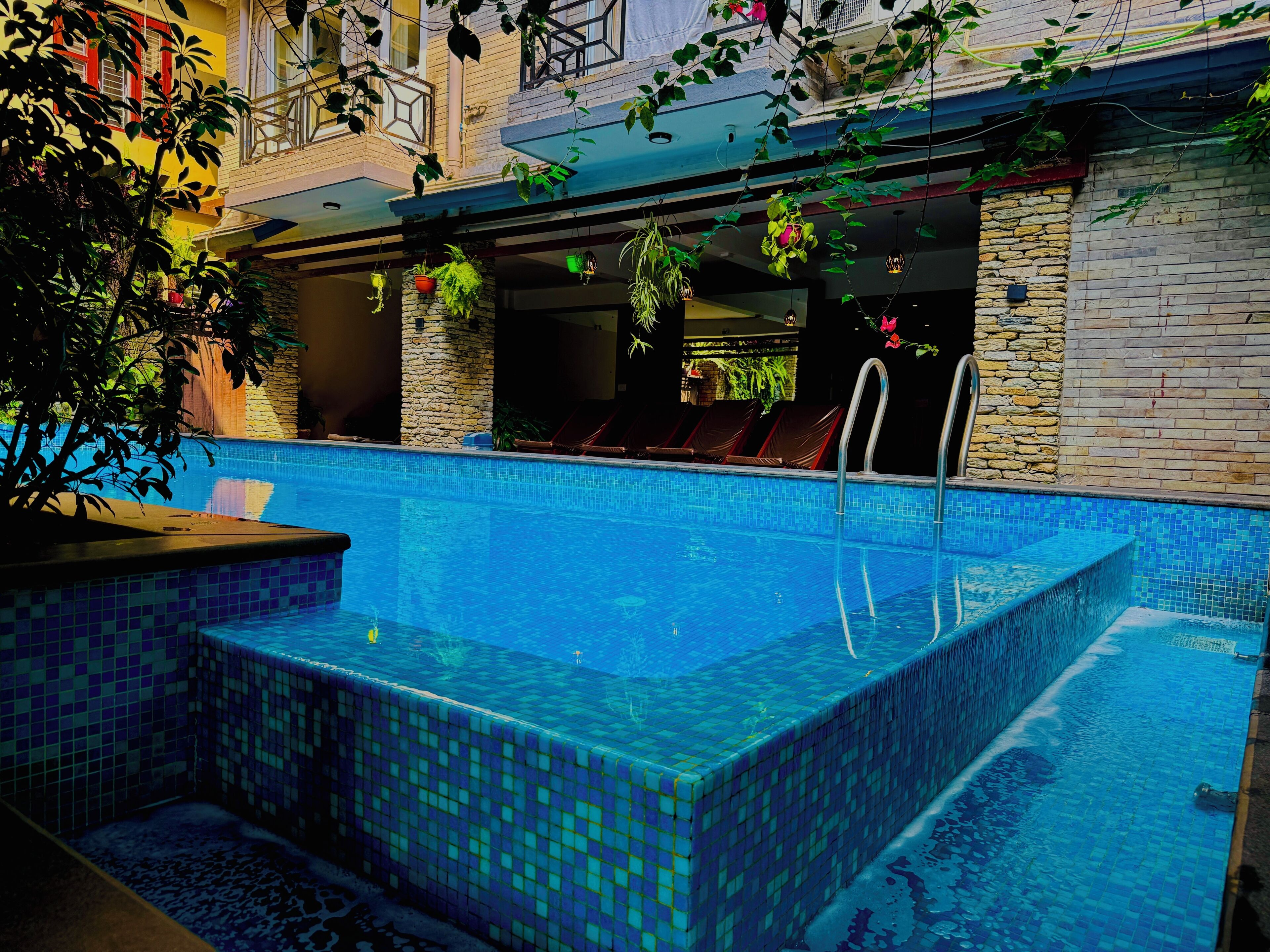 A rectangular pool with blue mosaic tiles, surrounded by a stone wall and a balcony with red cushions.