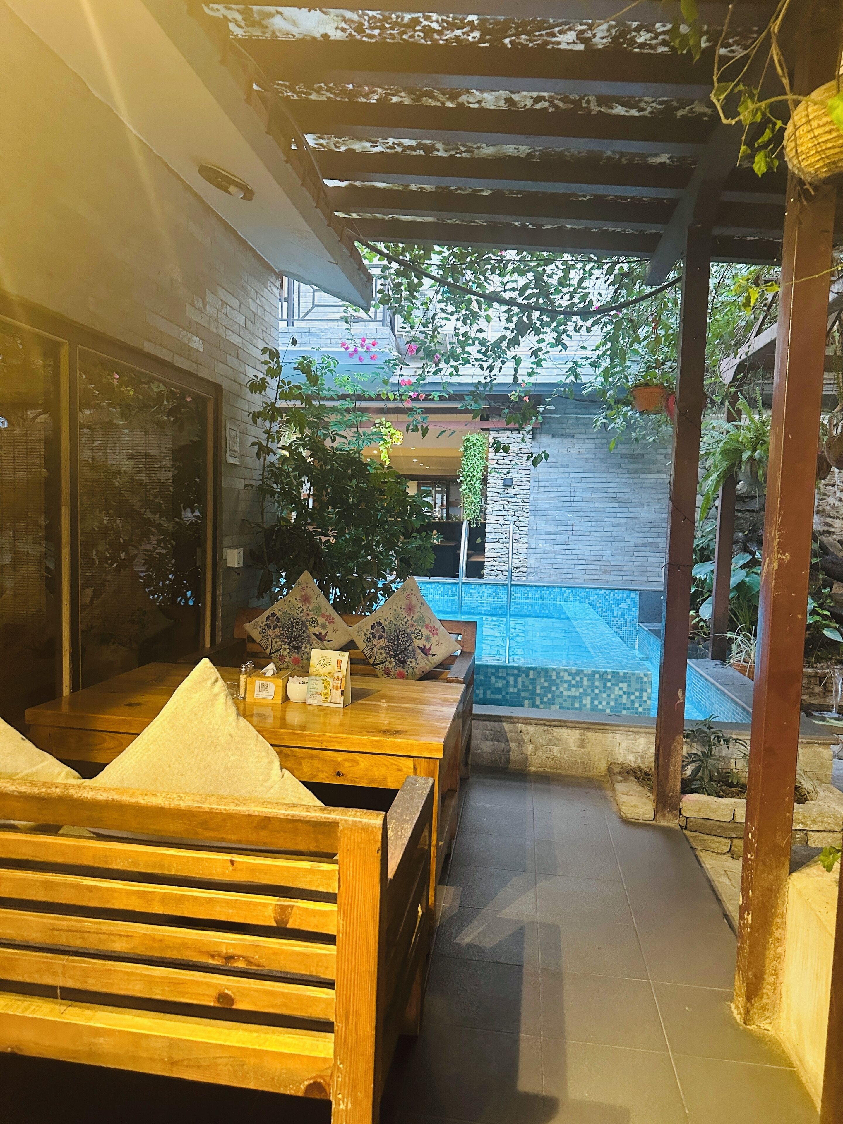A wooden bench and table set up under a pergola with a view of a swimming pool and greenery in the background.