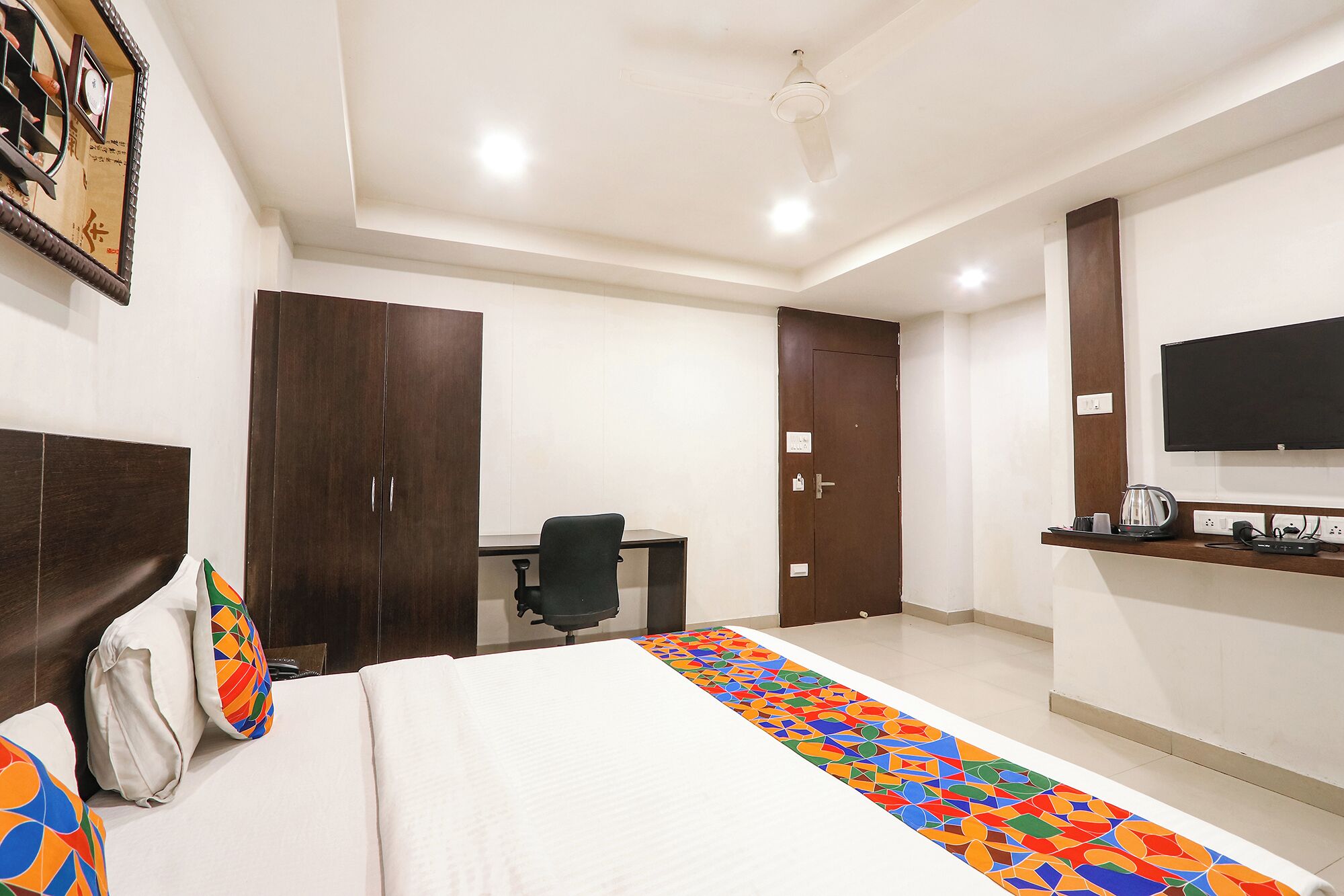 A hotel room with a bed, a desk, a chair, a wardrobe, a television, and a power strip.