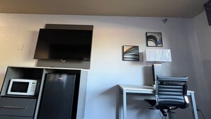 Habitación con televisor de pantalla plana montado en la pared, microondas y refrigerador en un mueble empotrado. Incluye una silla y un escritorio con lámpara.