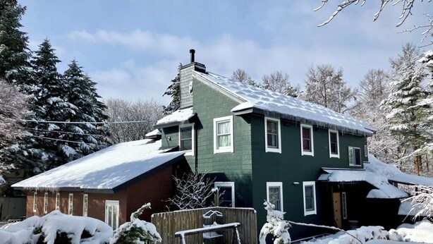 A two-story house with a green exterior and white trim, surrounded by snow-covered trees and a wooden fence.