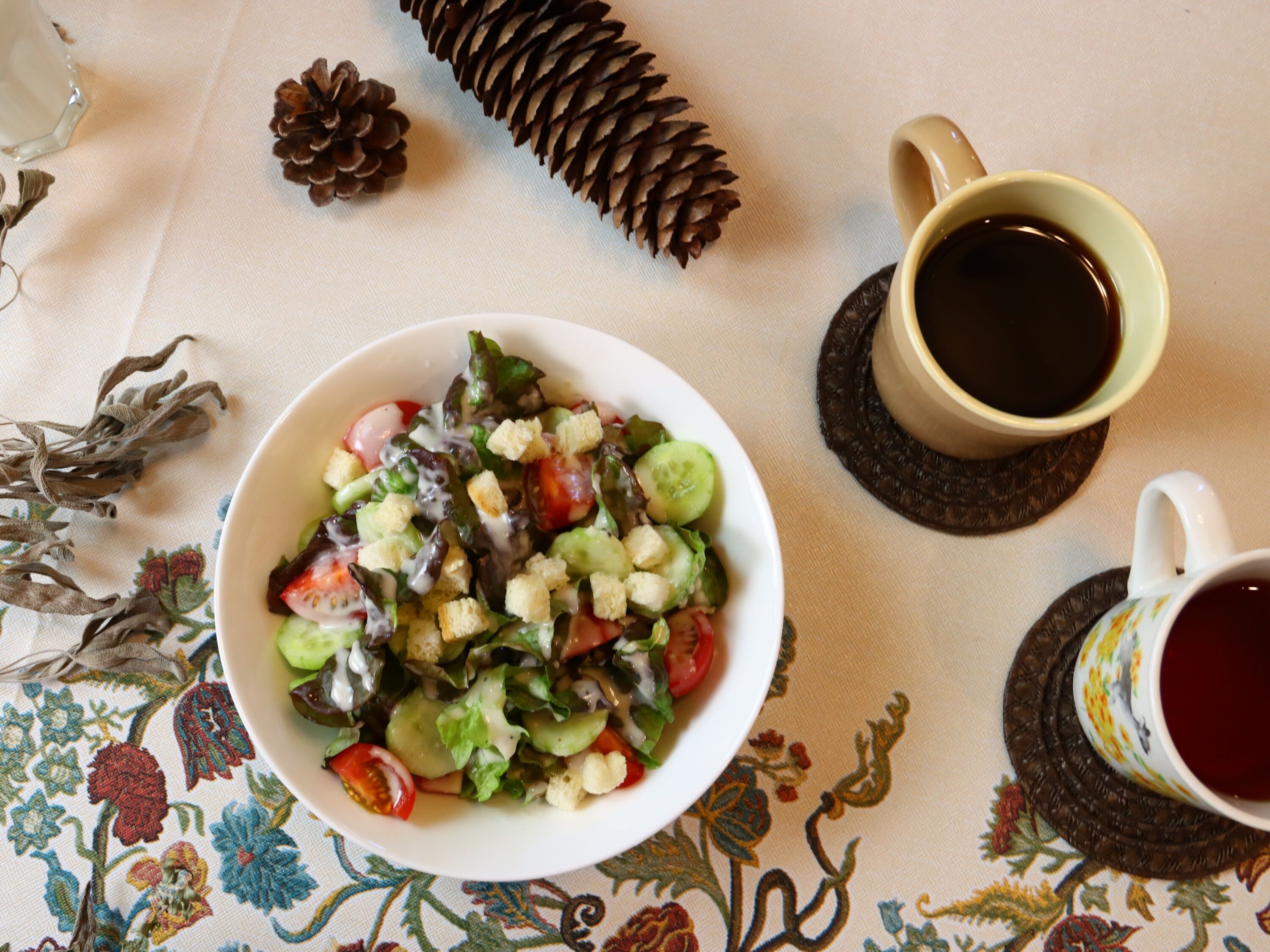 A table with a bowl of salad, two cups of coffee, and decorative pine cones.