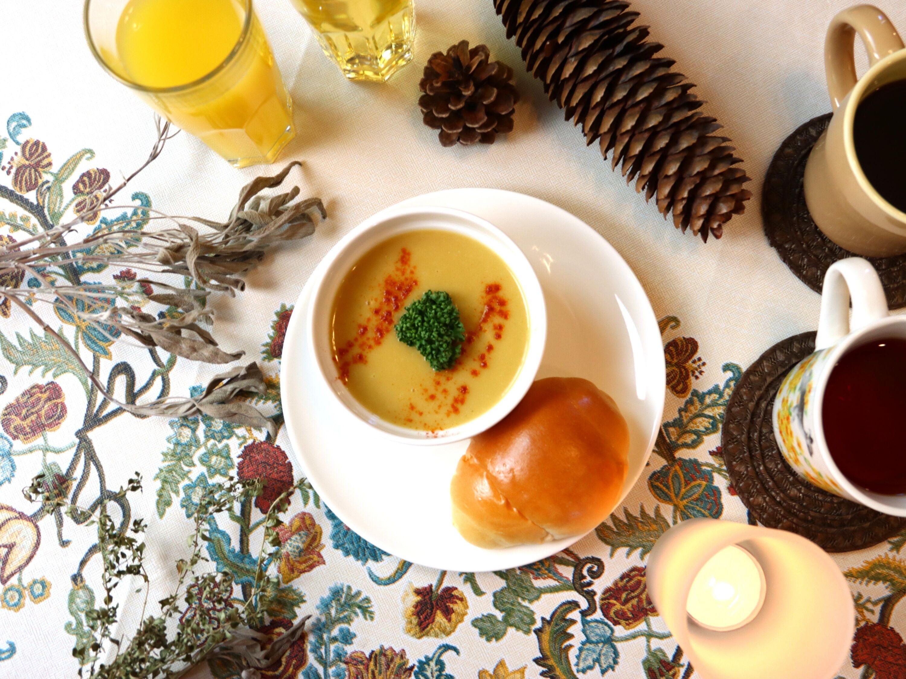 A table setting with a bowl of soup, a bread roll, a glass of juice, a candle, and a cup of tea.