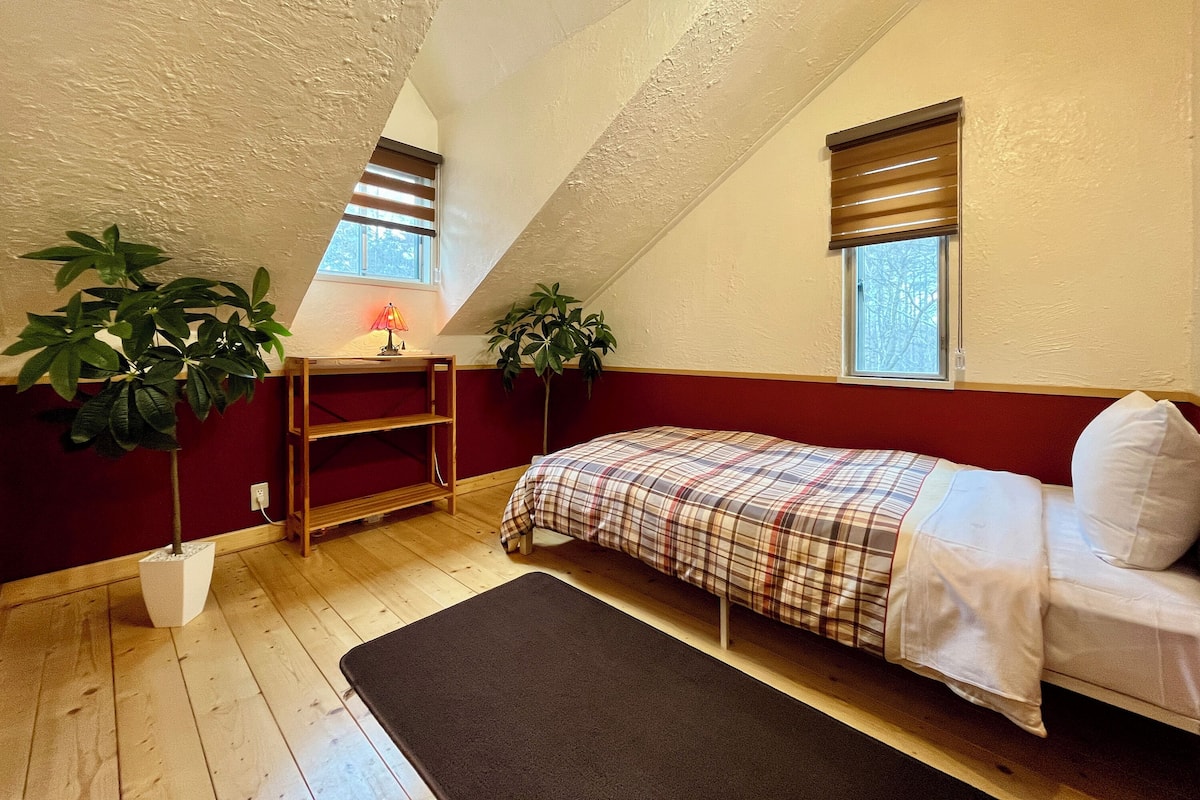 Una habitación pequeña y acogedora con techo inclinado, una cama con colcha escocesa, un escritorio con lámpara y dos plantas en macetas.