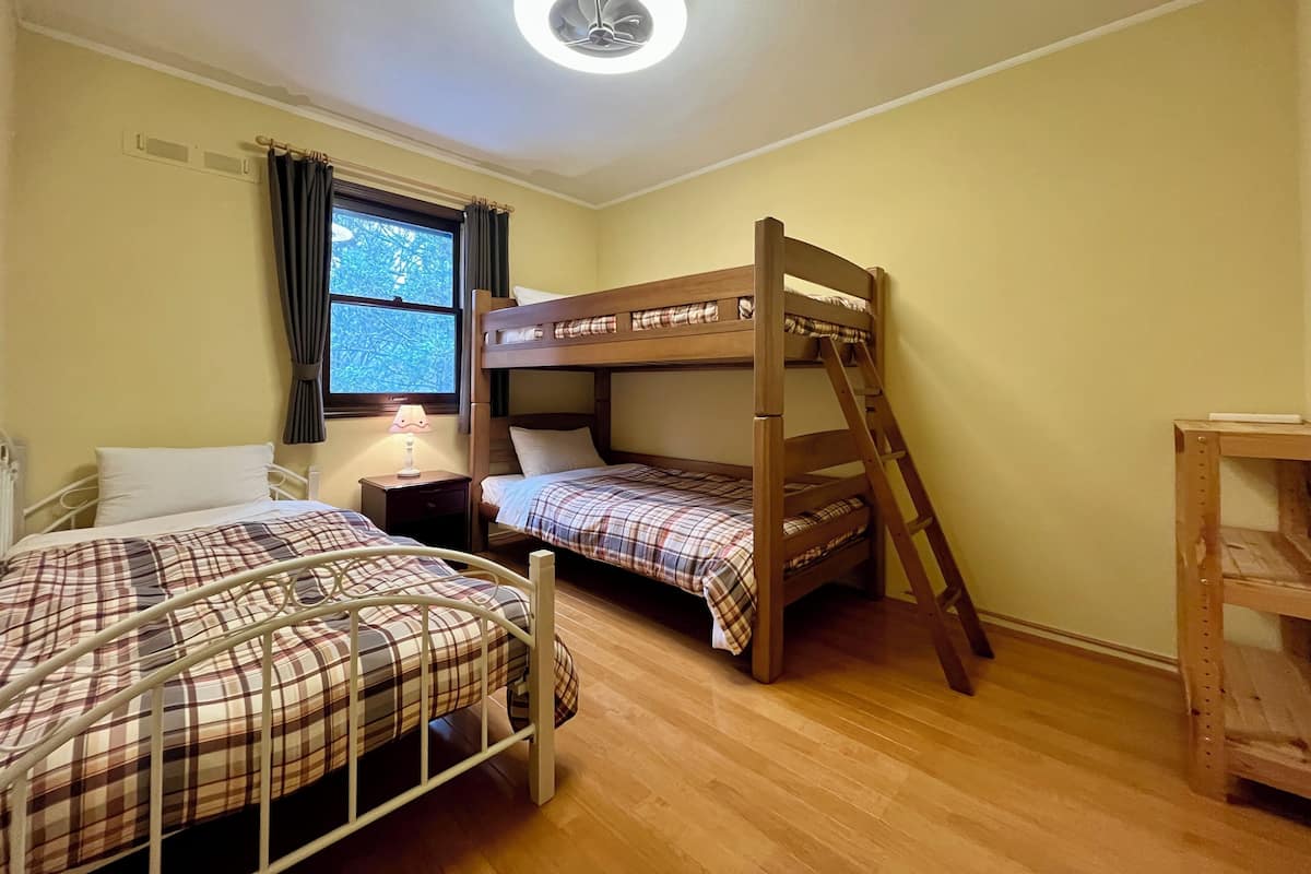 Habitación con dos camas, una literas y otra individual, ambas con ropa de cama de cuadros. Se ve una ventana con cortinas, una mesita de noche con lámpara y una lámpara en el techo.