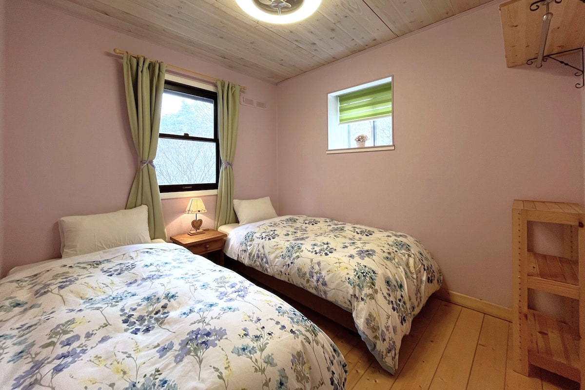 Un dormitorio con dos camas, una ventana con cortinas, una mesita de noche con lámpara y un ventilador de techo.