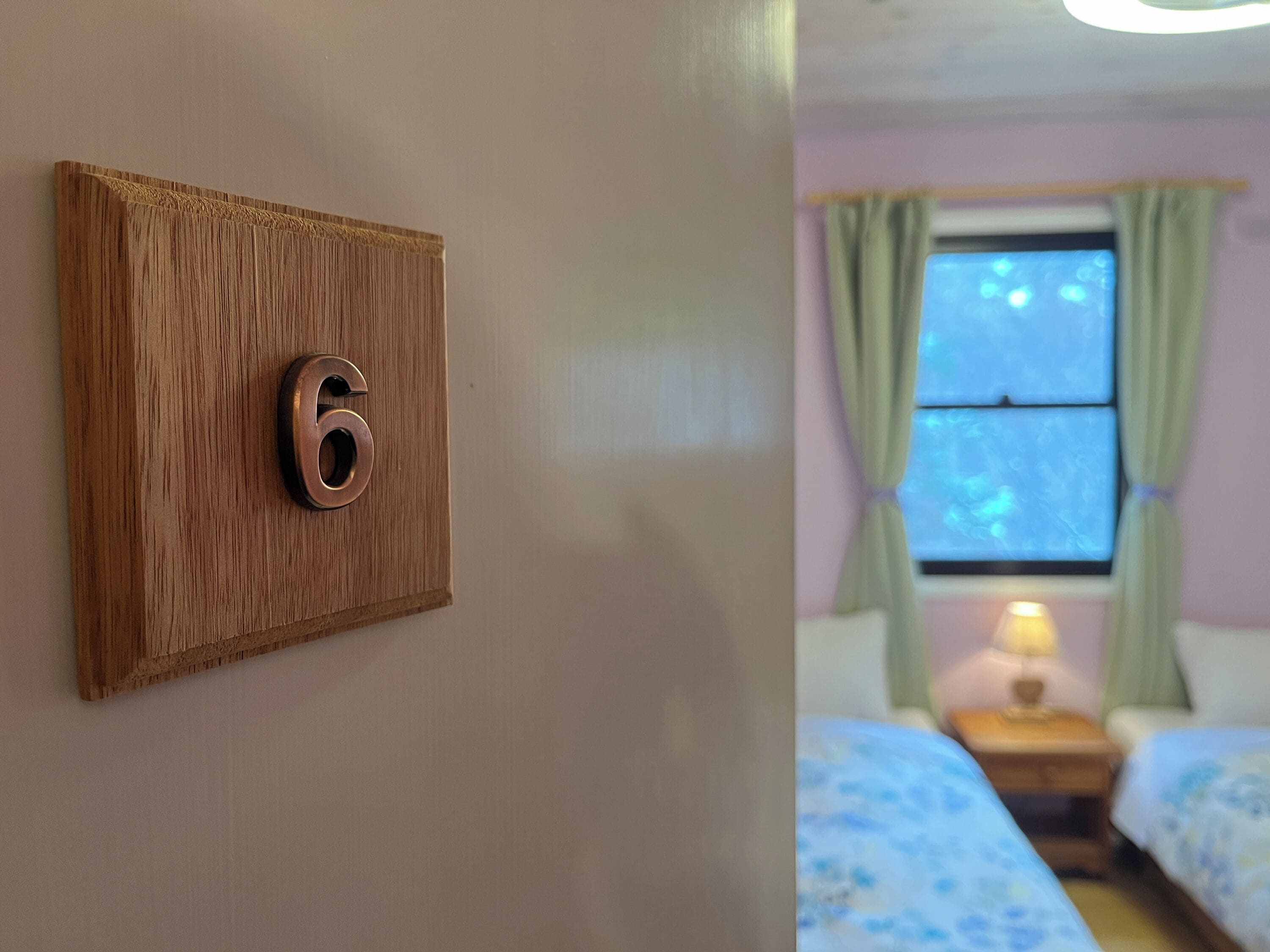 Una puerta de madera con un cartel con el número 6, una ventana con cortinas y una cama con una colcha estampada de flores.