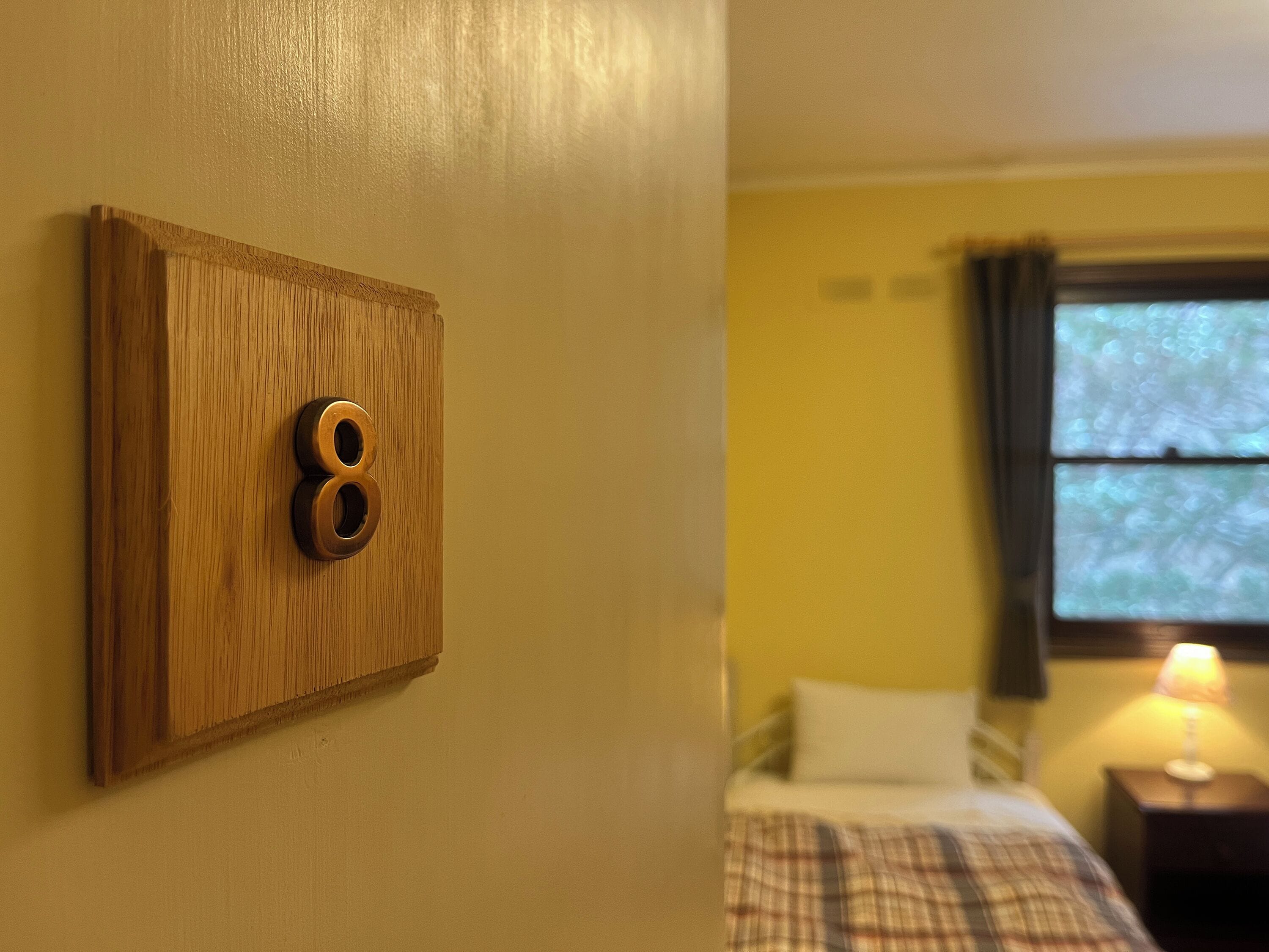 Una habitación con puerta de madera marcada con el número 8, una cama con manta escocesa, una mesita de noche con lámpara y una ventana con cortinas.