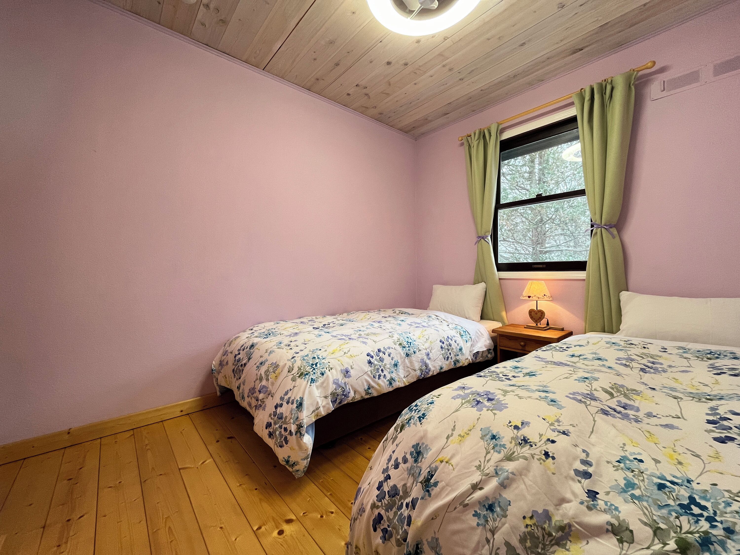 A bedroom with two beds, wooden flooring, a window with curtains, and a nightstand with a lamp.