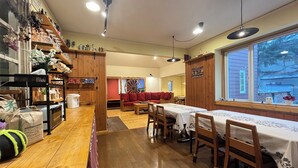 A cozy restaurant with wooden floors, a dining area with tables and chairs, a bar area with shelves and bottles, and a view through large windows.