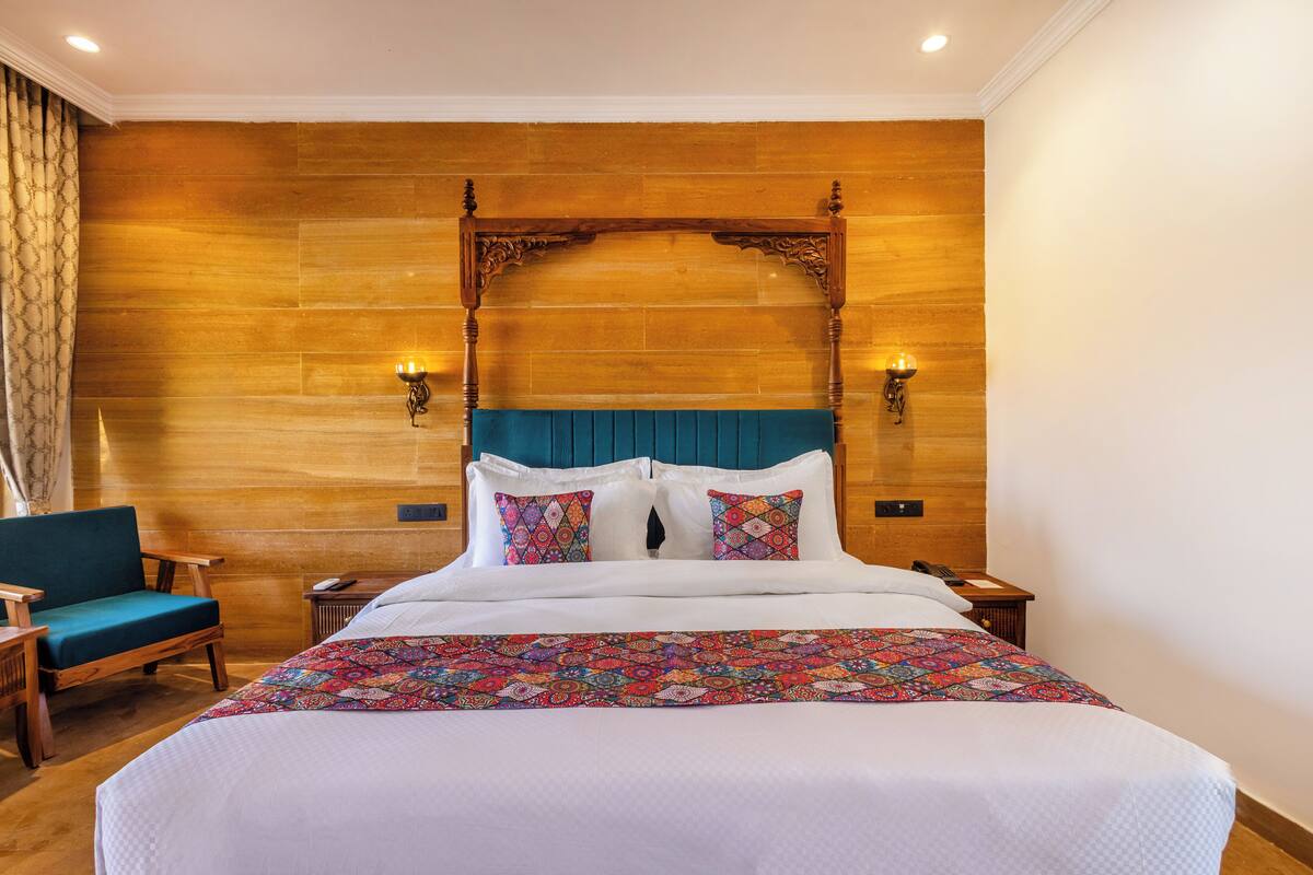 A hotel room with a large bed, a wooden headboard, two bedside tables with lamps, a chair, and a patterned bedspread.
