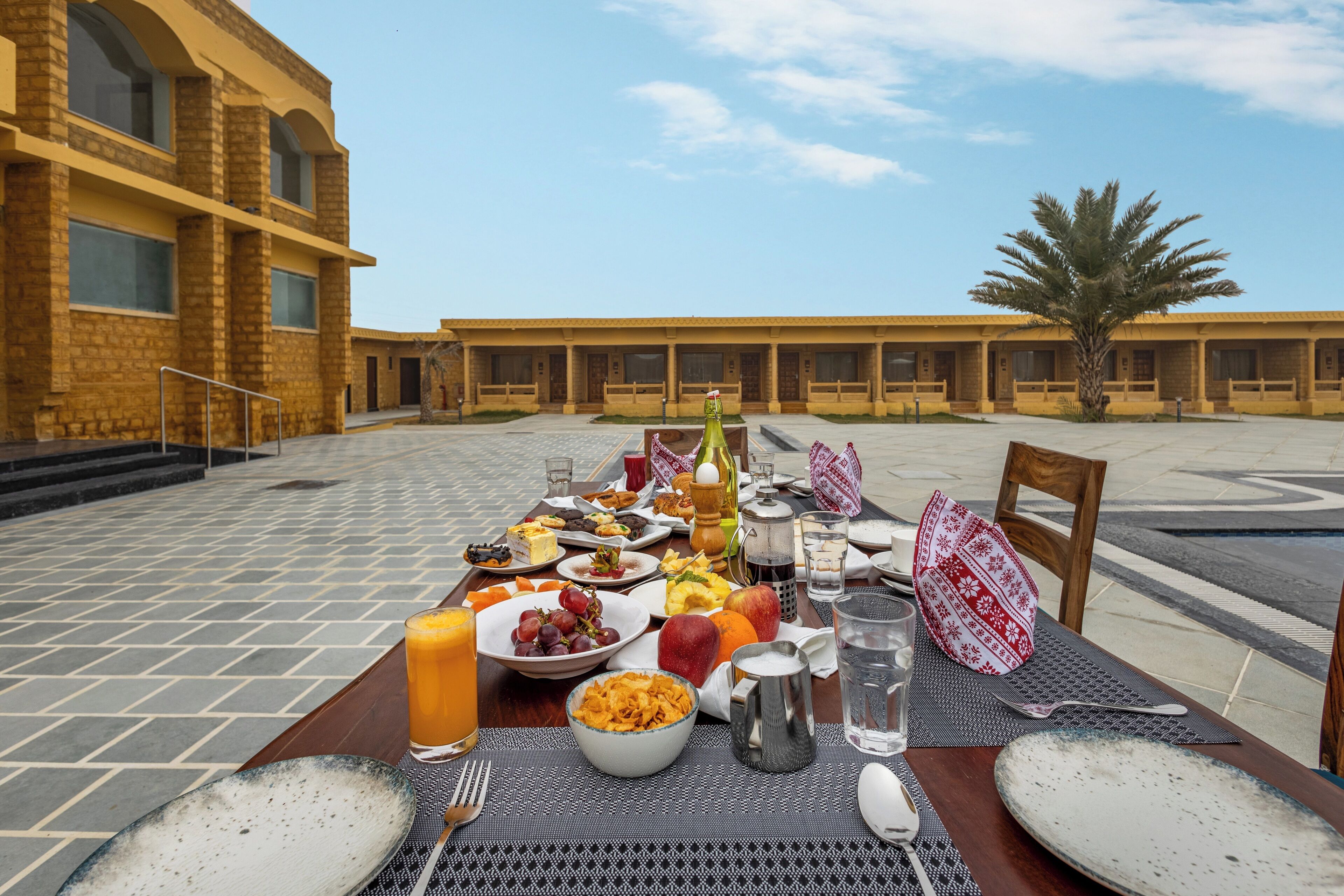 Una mesa dispuesta con alimentos para el desayuno, incluyendo frutas, un jarro de jugo y una jarra, en un patio al aire libre con un edificio al fondo.