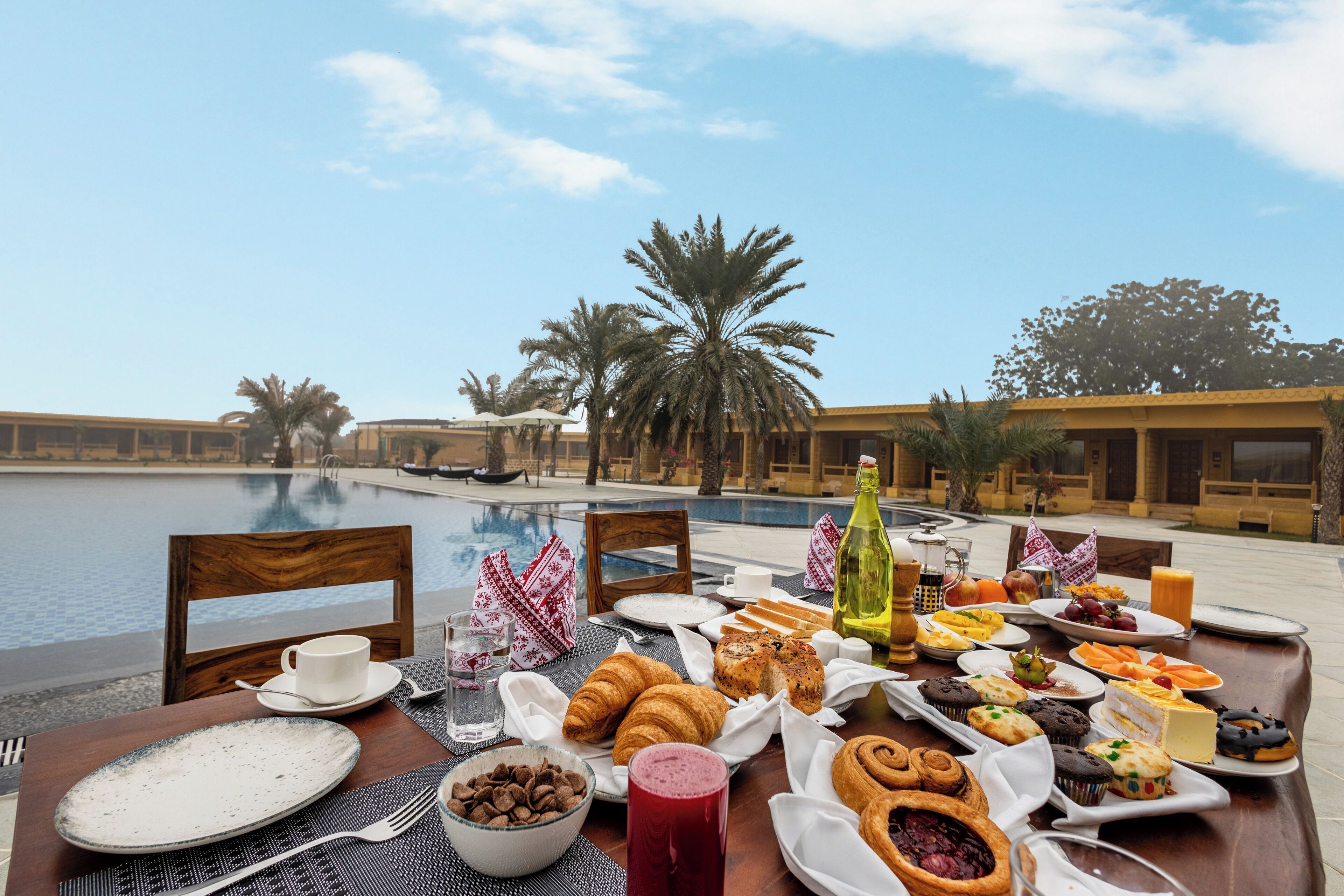 A breakfast table with croissants, pastries, and fruits by a poolside with palm trees and a clear sky.
