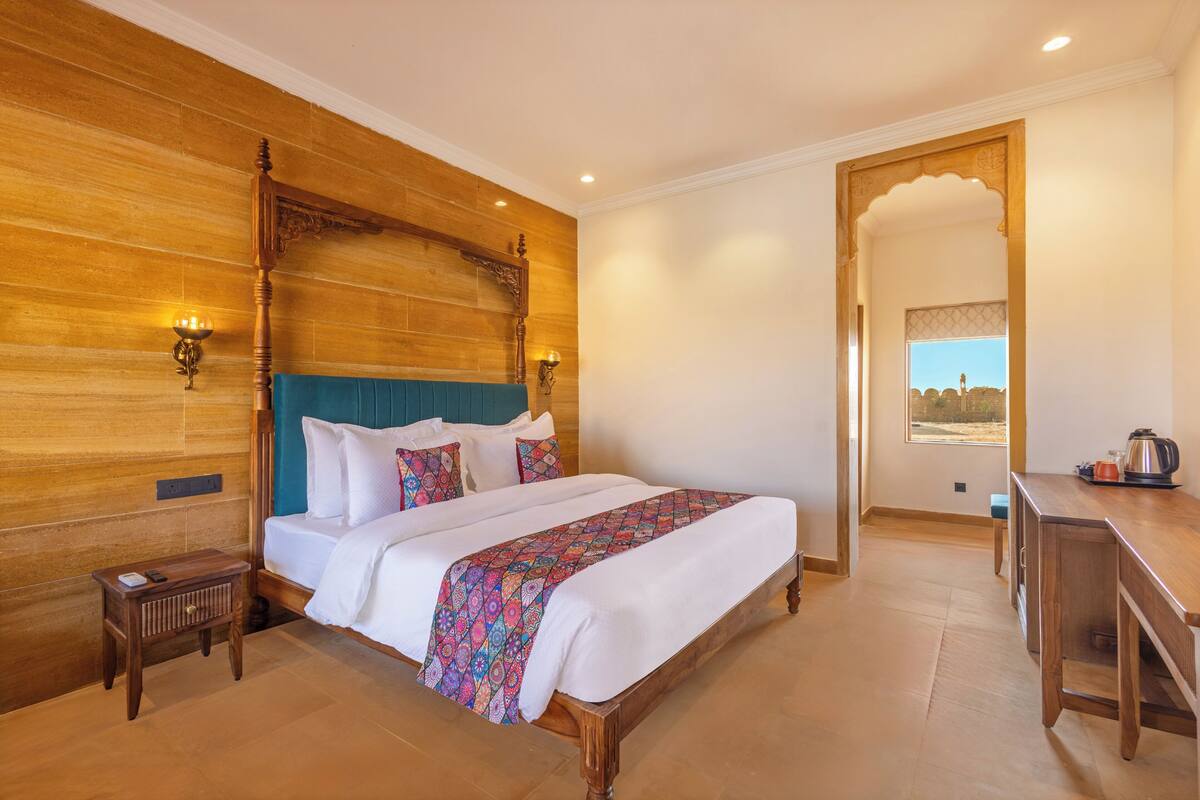 A hotel room with a wooden headboard, a large bed with white linens, a colorful bedspread, a small wooden nightstand, a desk with a kettle, and a view through an archway.