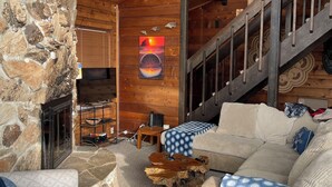 A cozy cabin interior with a stone fireplace, wooden ceiling, and a sofa with patterned cushions.