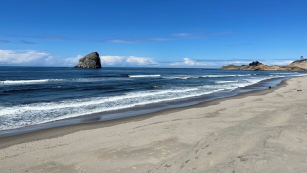 A beach with a large rock formation in the ocean, sandy shore, and distant hills.
