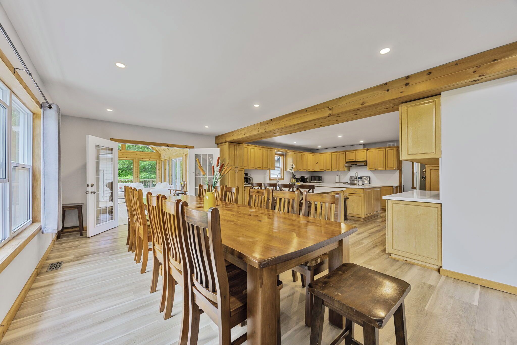 A spacious dining area with a large wooden table and chairs, a kitchen with wooden cabinets, and a view of greenery through an open door.
