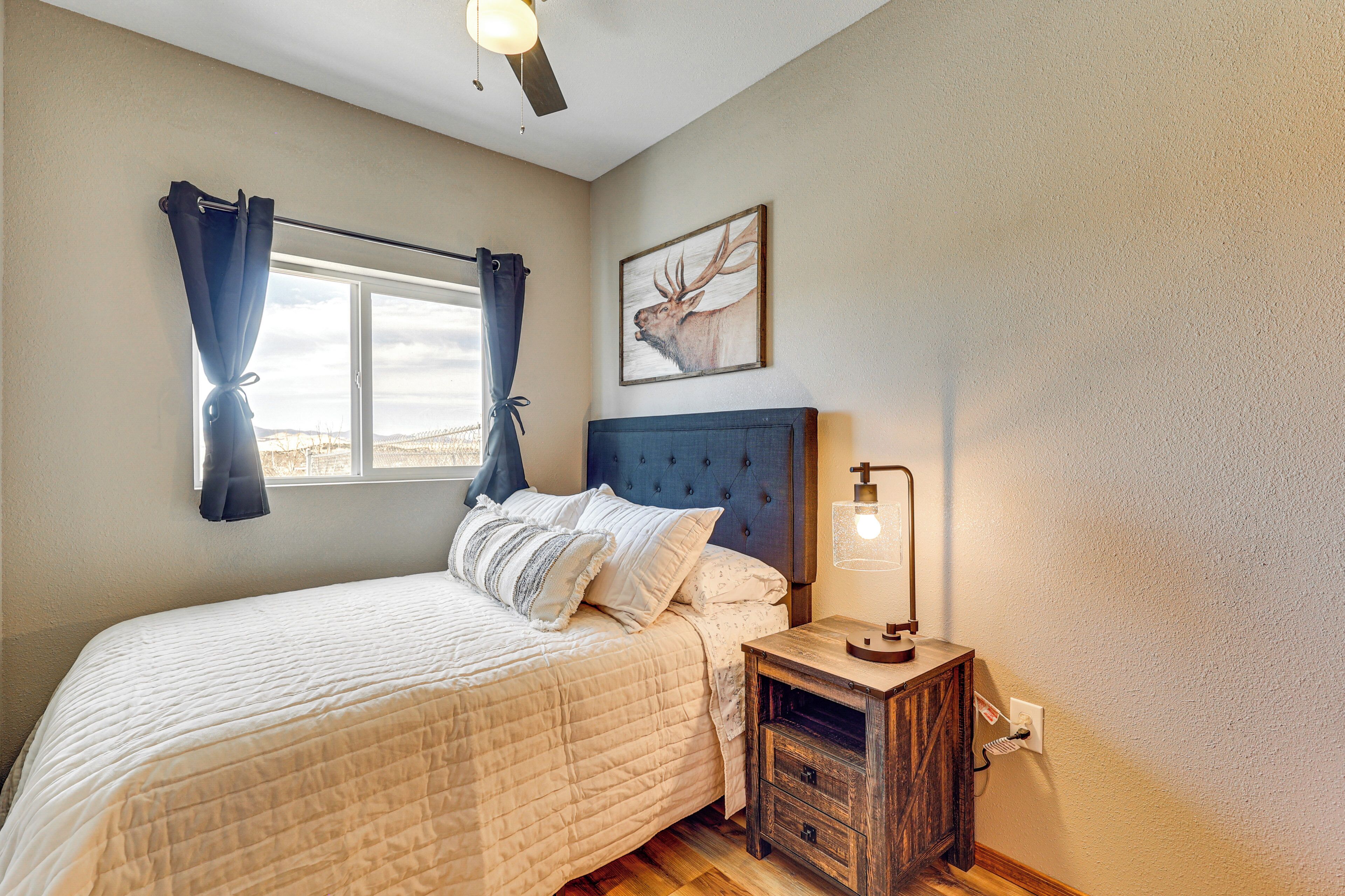 Un dormitorio con cama, mesita de noche, lámpara de pared, ventana con cortinas y un cuadro de un ciervo en la pared.