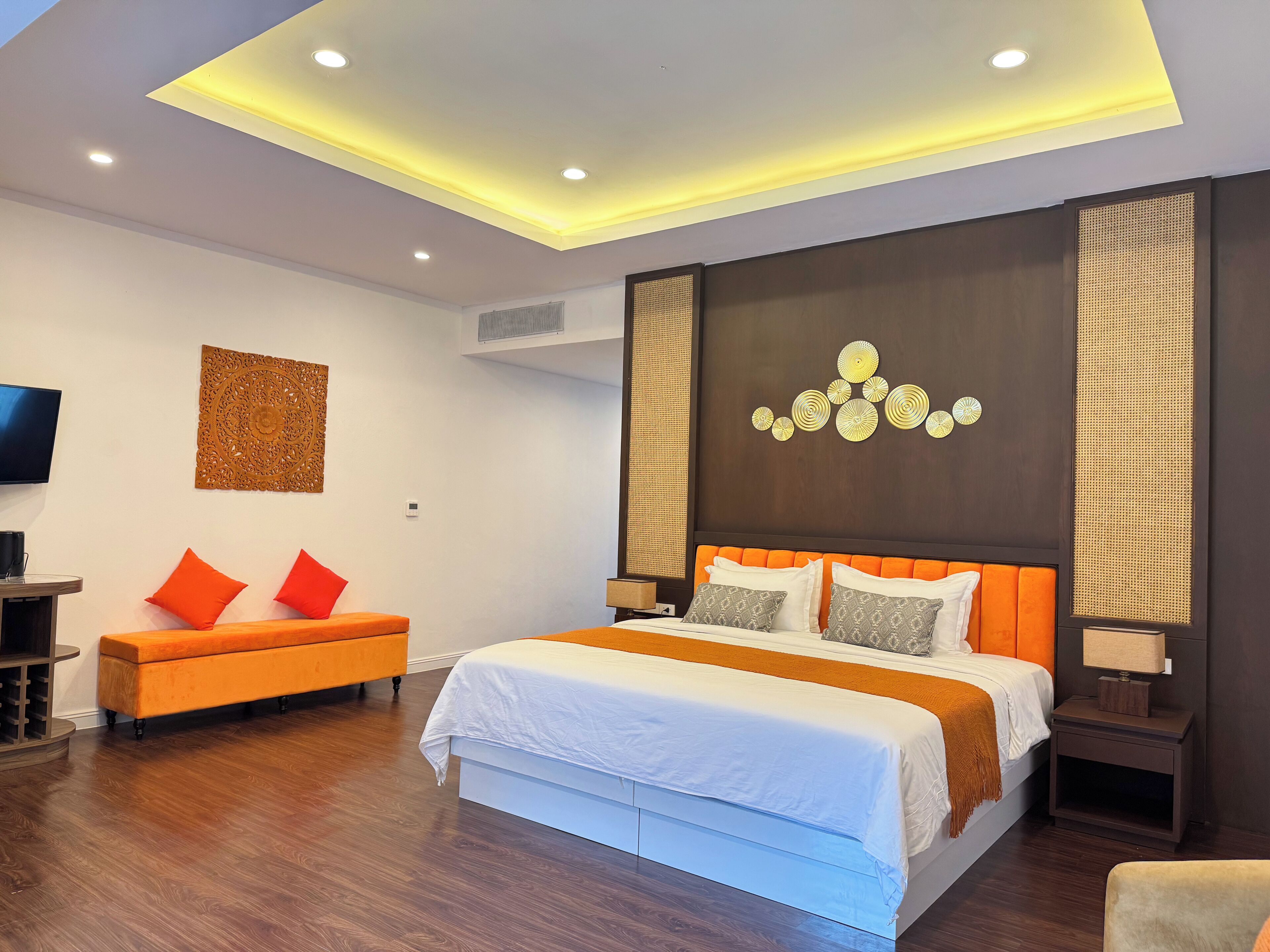 A modern hotel room with a large bed, orange accent pillows, a wooden bench, and a ceiling with recessed lighting.