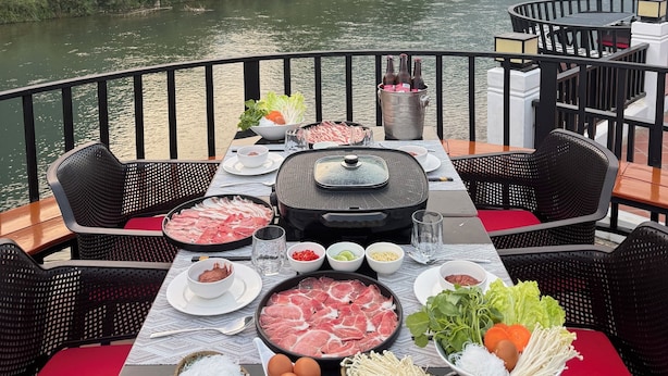 Ein Esstisch ist reichhaltig mit Speisen und einem heißen Grill gedeckt, mit Blick auf einen Fluss und Berge im Hintergrund.