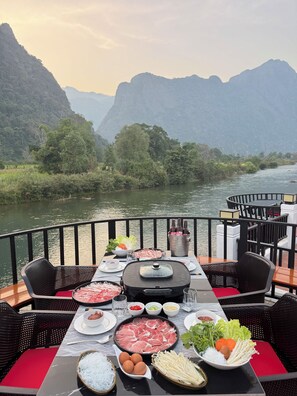 A dining table set with various food items and a hot grill, overlooking a river with mountains in the background. - Phalai view hotel (Vang Vieng)