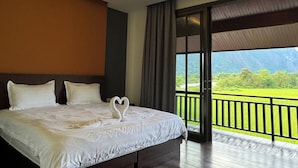 Ein Hotelzimmer mit einem großen Bett, Blick auf eine grüne Landschaft durch einen Balkon und einer Deckenleuchte.