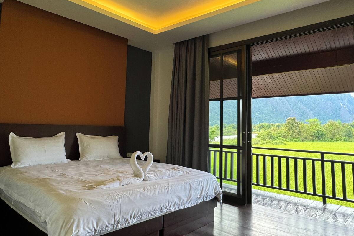 A hotel room with a large bed, a view of a green landscape through a balcony, and a ceiling light fixture.
