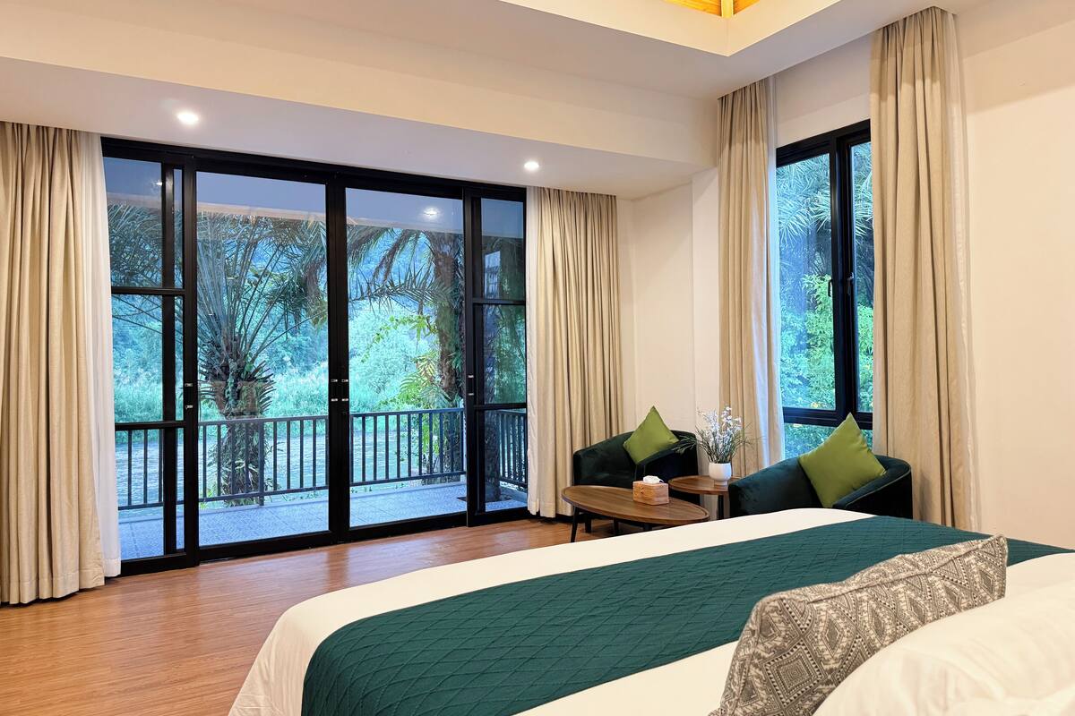 A spacious bedroom with a large bed, a small table, and a chair. It features large windows with a view of a lush garden.