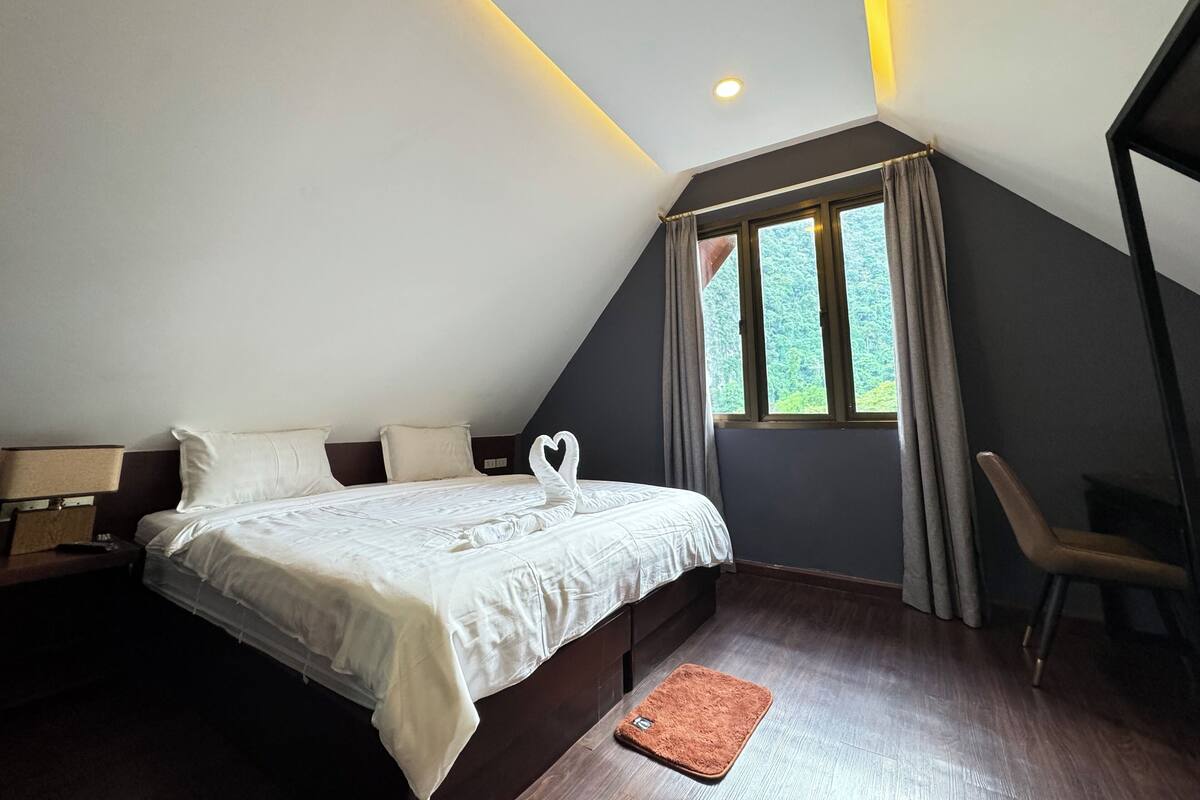 A bedroom with a bed, a nightstand, a chair, a window with curtains, and a ceiling light.