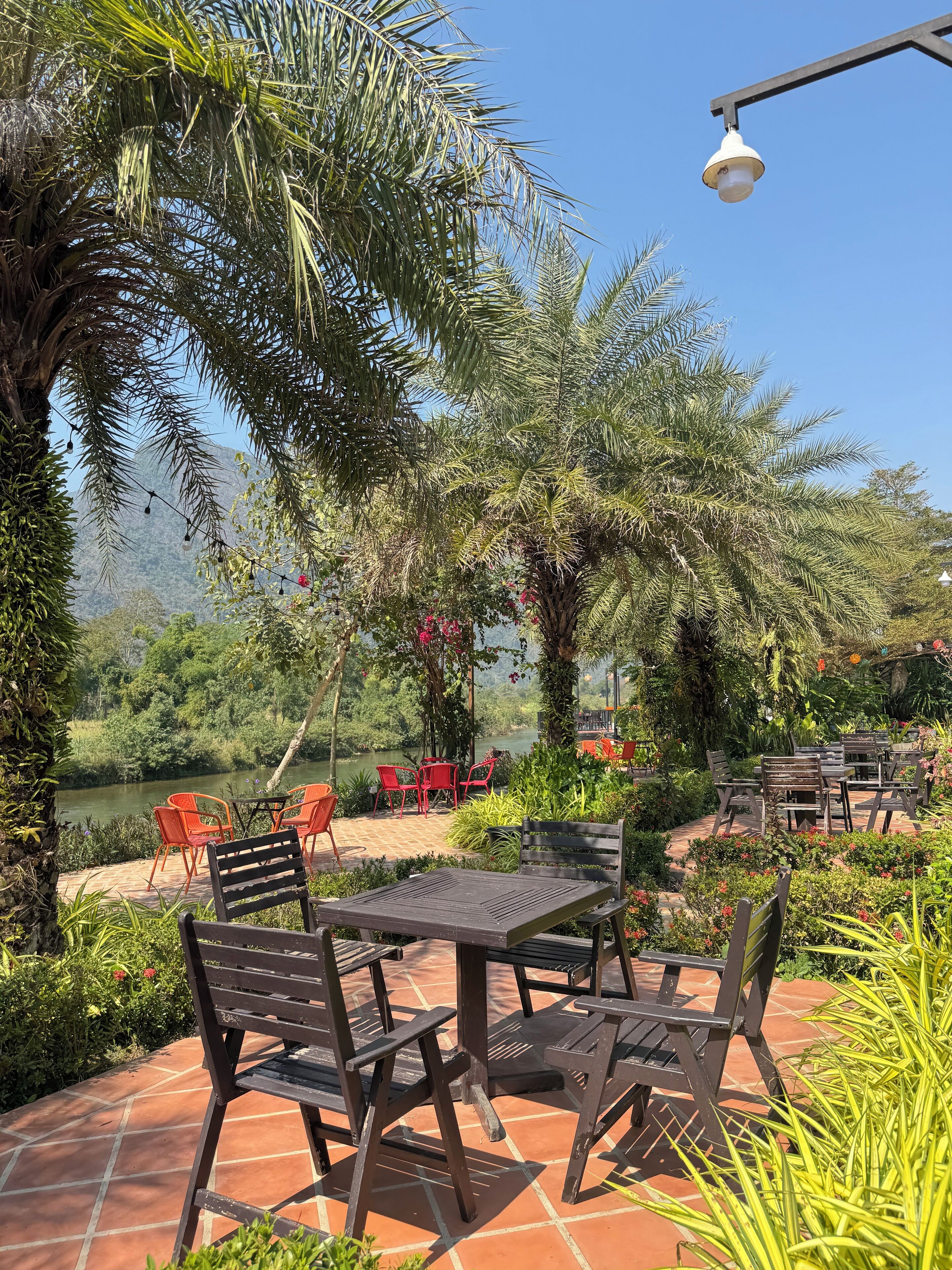An outdoor seating area with tables and chairs, surrounded by lush greenery and a view of a river.