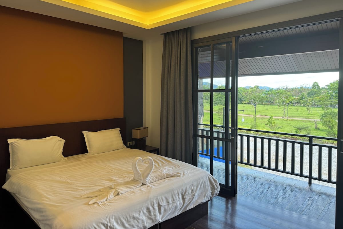 A hotel room with a large bed, a nightstand, a lamp, and a view of a green landscape through a large window.