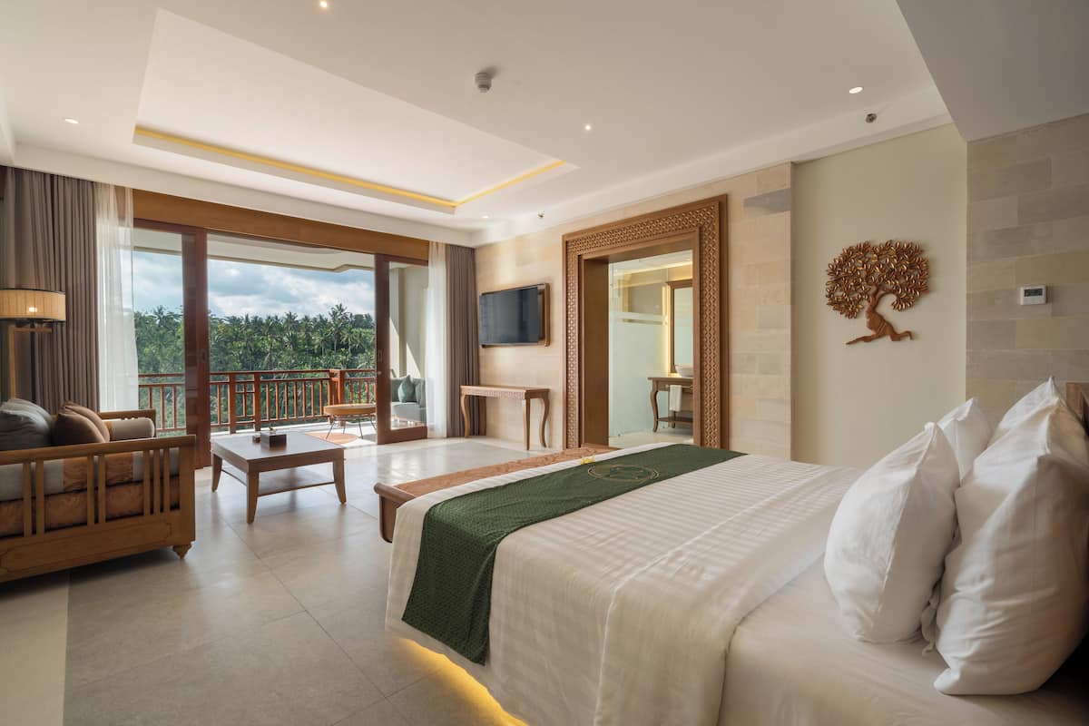 A modern hotel room with a large bed, a sofa, a desk, and a balcony with a view of the greenery.