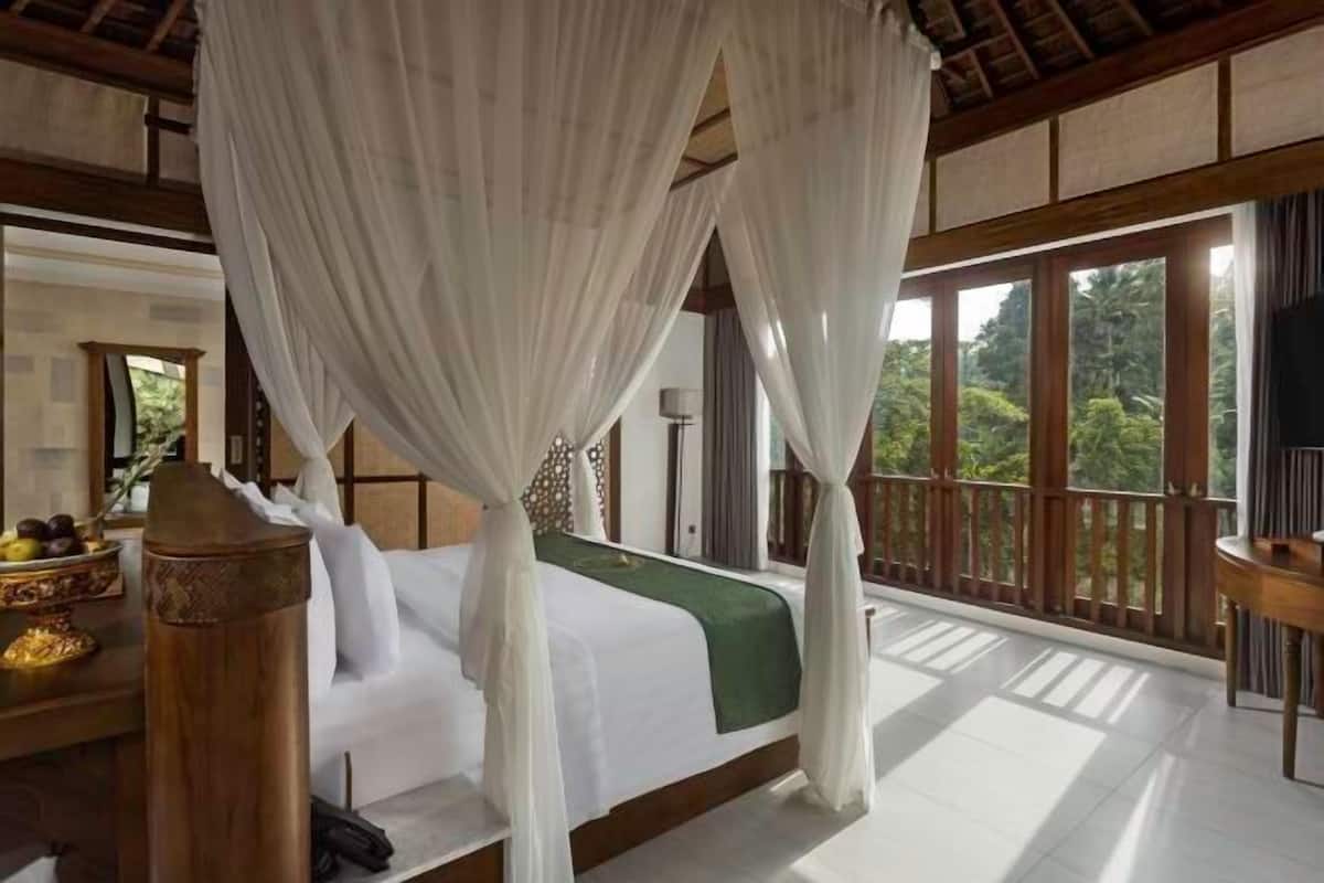 A room with a bed under a canopy, a wooden desk with a lamp, and a view of greenery outside.