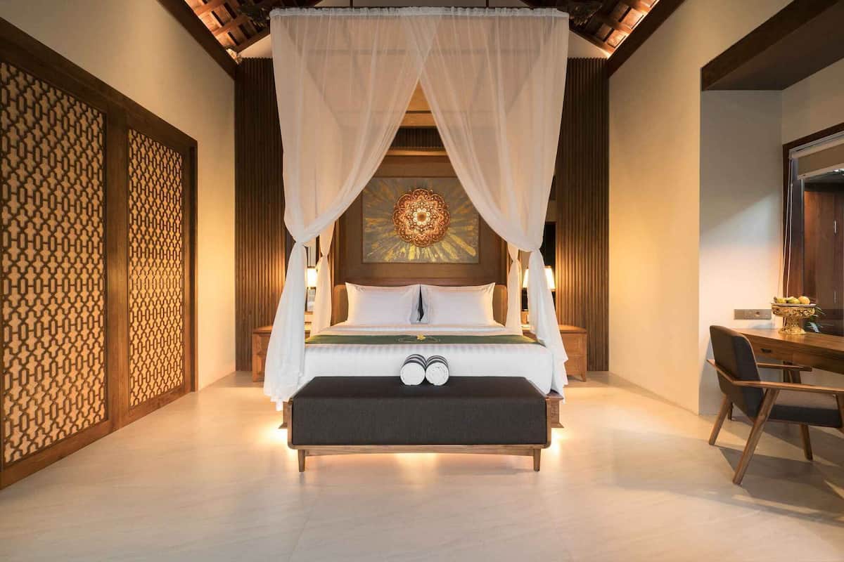 A four-poster canopy bed with a decorative headboard, a bench at the foot of the bed, and a wooden desk with a chair to the right.