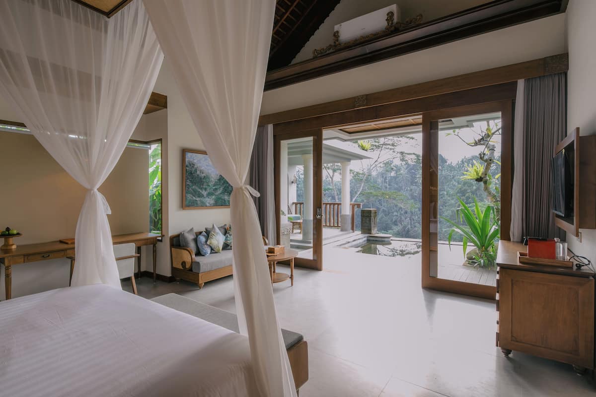 A bedroom with a large bed, a desk, a sofa, and a view of a garden through large sliding doors.