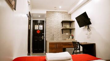 A hotel room with a bed, a desk, a chair, a TV mounted on the wall, and a door with a red circle sticker.