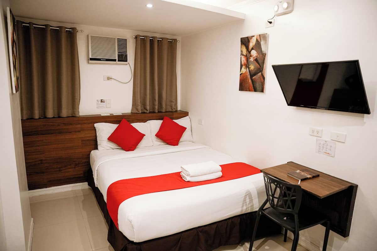 Quarto de hotel com cama, escrivaninha, cadeira, televisão e cortinas.